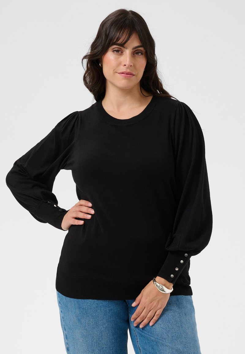 Femme aux longs cheveux foncés porte un haut noir à manches longues avec manches bouffantes et poignets à boutons, assorti à un jean bleu, debout avec assurance.
