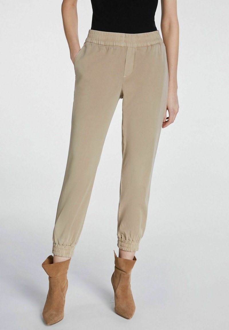 Pantalon beige à taille élastique avec poches latérales et chevilles resserrées, associé à des bottines beige. Tissu lisse avec une coupe décontractée.