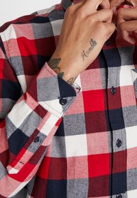 Camisa de franela a cuadros en rojo, azul marino y blanco, con mangas largas, puños con botones y cuello estándar. Cuenta con botones oscuros y un bolsillo en el pecho.