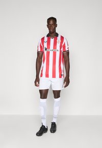 Rot-weiße gestreifte Fußballtrikot mit schwarzem Kragen, weißen Shorts, schwarzen Fußballschuhen und kniehohen weißen Socken mit strukturierten Details.