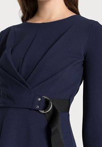 Mörkblå klänning med strukturerad inslagning, med en metallring och svart remaccent. Slät tyg med subtil textur.