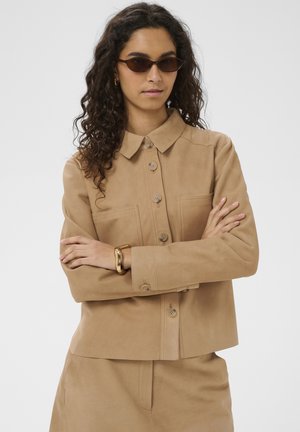 Femme aux cheveux bouclés portant des lunettes de soleil marron, une veste à boutons beige avec poches poitrine, un pantalon assorti et un bracelet épais en or, les bras croisés.