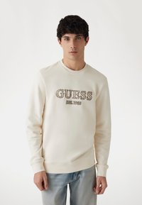 Felpa color crema con ricamo decorativo "GUESS EST. 1981" sul petto, con polsini e vita a coste. Indossata con jeans blu.