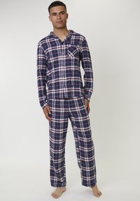 Joven de pie descalzo, vistiendo un conjunto de pijama de manga larga en cuadros azul marino, blanco y rojo, frente a un fondo liso.