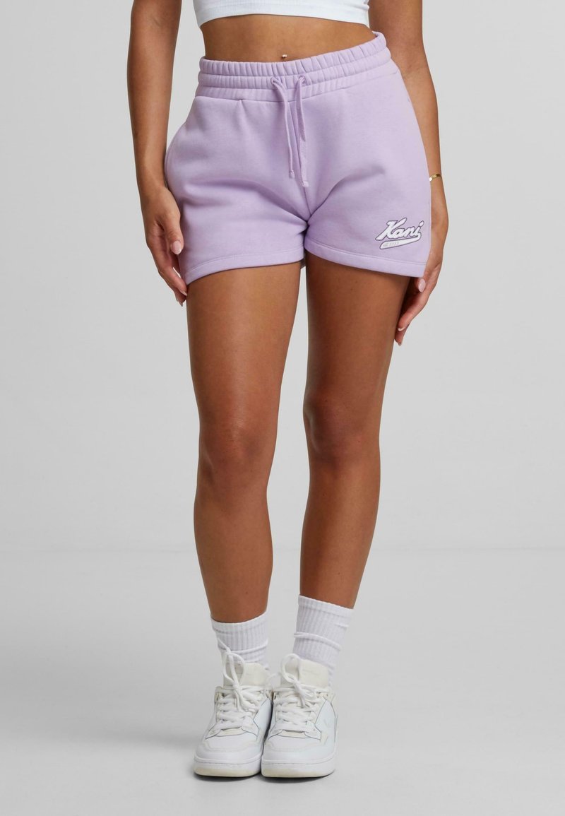 Personne portant un short violet clair à cordon, un crop top blanc, des chaussettes blanches et des baskets blanches, debout devant un fond uni.