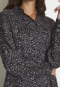 Chemise noire à boutons avec un imprimé léopard beige et blanc dispersé, dotée d'un col volanté et de manches longues. Texture de tissu douce.