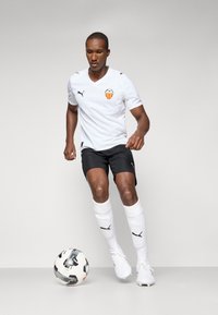 Hvit fotballtrøye med logo, svarte shorts og hvite sokker. Spiller i bevegelse, holder en fotball; minimalistisk bakgrunn.
