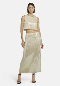 Glitzerndes Zweiteiler-Set mit einem kurzen Oberteil und einem langen Rock. Hellbeige Farbe, glänzende Textur, Seitenschlitz am Rock und schlichte Träger.