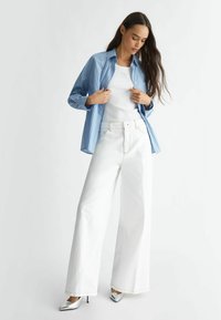 Camisa de botones a rayas azules sobre una camiseta blanca, combinada con jeans anchos blancos y tacones plateados de punta en un fondo claro.