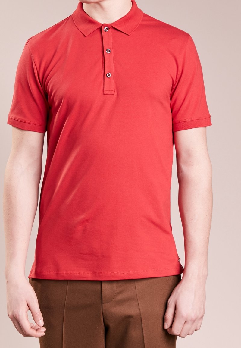 Homme portant un polo rouge à manches courtes avec trois boutons et un pantalon marron sur un fond uni.