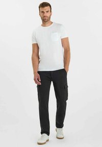 Witte t-shirt met korte mouwen en een borstzak, gecombineerd met zwarte cargobroek en witte sneakers met bruine zolen, gedragen door een mannelijke model.