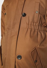 Chaqueta exterior marrón con una textura suave y mate; presenta botones negros, una cinturilla elástica y un bolsillo lateral.