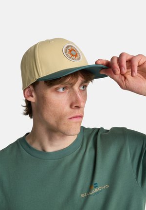 Beige Kappe mit einer dunkelgrünen Krempe und einem runden Billabong-Logo mit einem Sonnenmotiv. Der Stoff wirkt glatt und die Passform ist strukturiert.