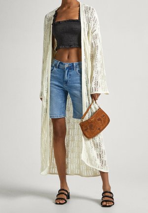 Femme portant un long cardigan blanc en maille ouverte, un top court noir tube, un short en jean, des sandales noires, et tenant un petit sac à main marron.