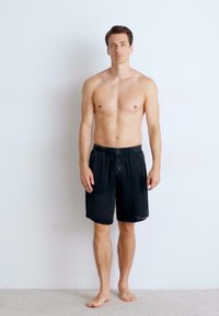 Black athletic shorts with a button closure, featuring a lightweight, smooth texture. The model stands barefoot against a plain background.