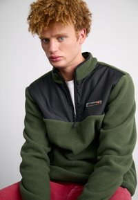 Green fleece pullover with a black nylon upper section, half-zip design, and a logo patch. Soft texture and relaxed fit, paired with pink pants.