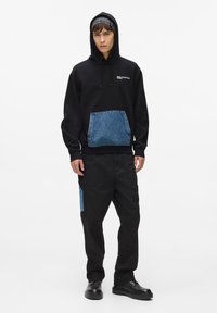 Juoda maikutė su džinsiniu kišeniu, turinti raištelio kapšą ir logotipą. Derinama su juodomis kelnėmis, kuriose taip pat yra džinsinis akcento kišenė.