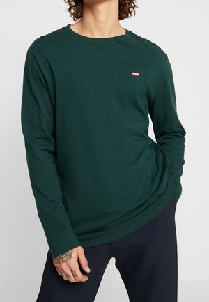 Langærmet mørkegrøn bomulds-T-shirt med rund halsudskæring og lille rød logo-patch på brystet, kombineret med mørke bukser.