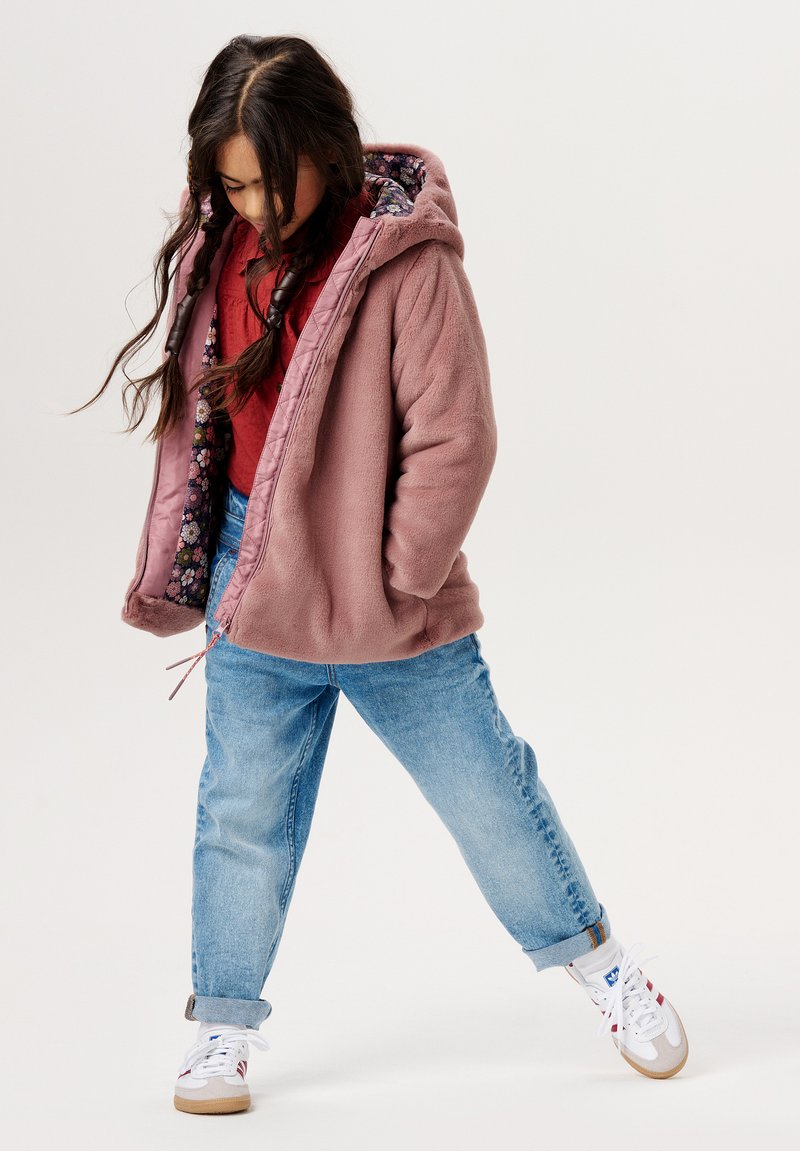 Roze fleecejack met een bloemenvoering, gecombineerd met lichtblauwe spijkerbroek en witte sneakers. Het jack heeft een capuchon en een rits.