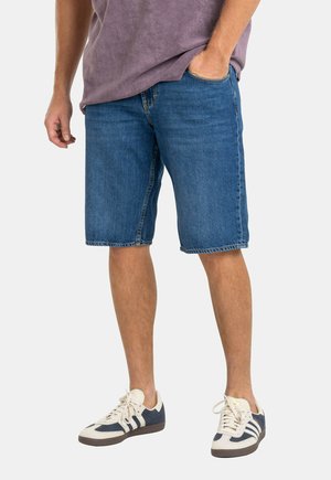 Mann trägt blaue Denim-Shorts, ein lila Hemd und weiße Sneaker mit dunkelblauen Streifen, steht mit einer Hand in der Tasche.