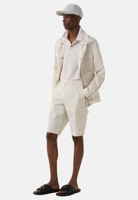 Beige bomuldsjakke med flere lommer, lyserød polo-shirt, beige cargo-shorts og sorte slid-sandaler, suppleret med en grå kasket.
