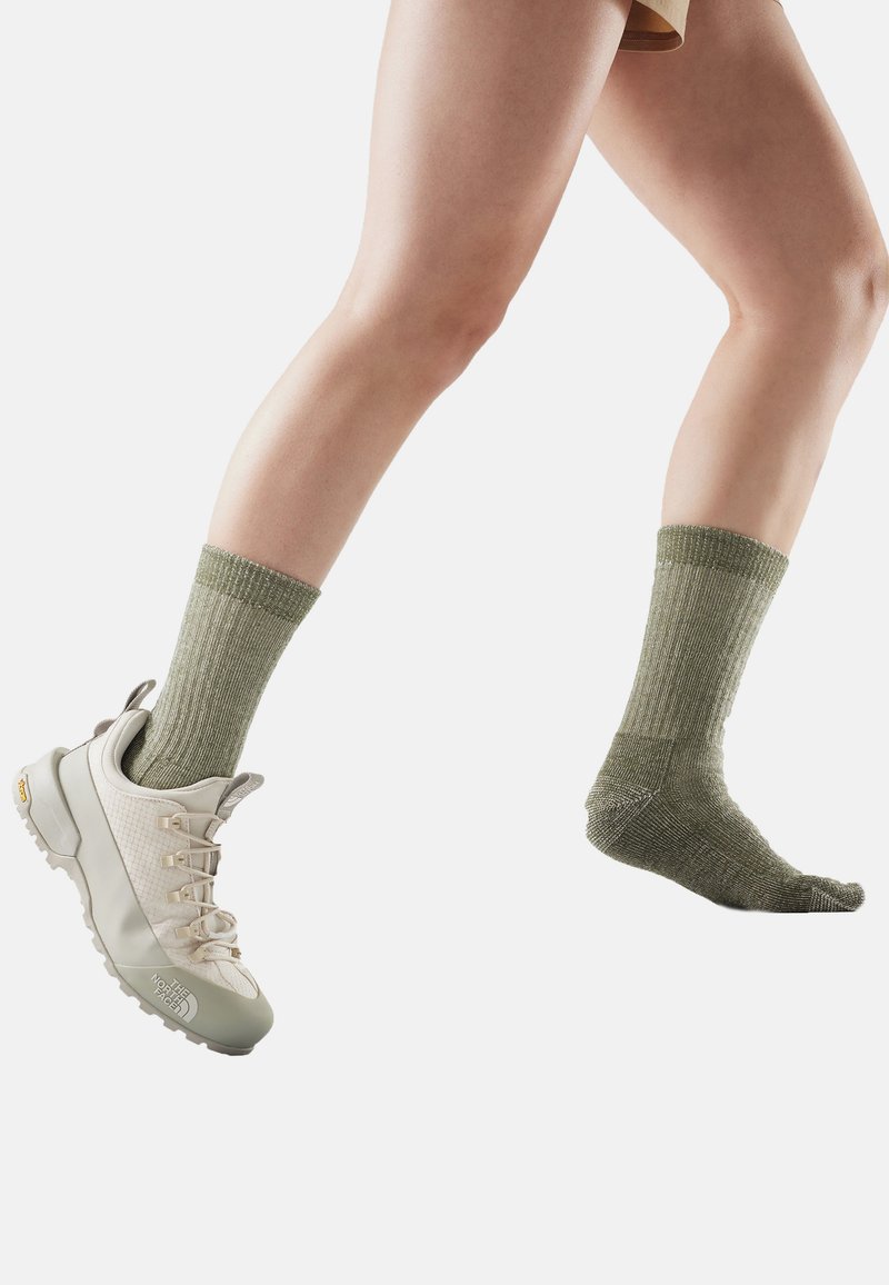 Gambe che camminano con una sneaker beige chiaro al piede sinistro e un calzino verde oliva al piede destro su sfondo bianco.
