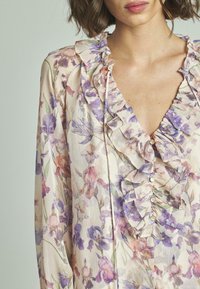 Blumenbluse mit Rüschenausschnitt, aus cremefarbigem Stoff mit lila und pinken Blumenmustern. Knöpfe entlang der Vorderseite mit weicher, strukturierter Oberfläche.