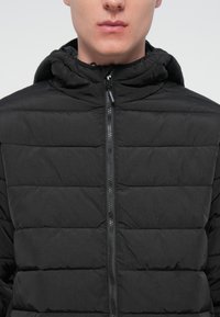 Veste rembourrée noire avec un col haut, motif matelassé horizontal, fermeture éclair frontale et capuche avec cordon de serrage. Texture lisse et design léger.