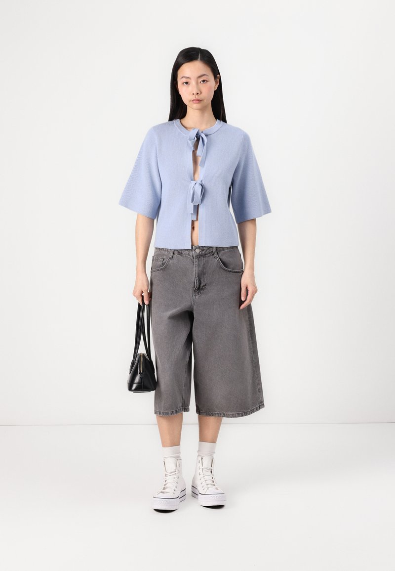 Jasnoniebieski, prążkowany krótki kardigan z wiązaniem z przodu, w połączeniu z luźnymi, szarymi dżinsowymi kulotami oraz białymi trampkami za kostkę. Do zestawu dołączona czarna torba.