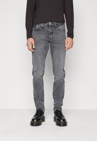 Jean en denim gris avec une coupe slim, design à cinq poches, texture délavée et coutures contrastantes, associé à des bottes noires à lacets.