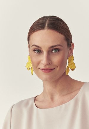 Žena so zelenými očami a jemným mejkapom, ktorá má na sebe veľké žlté náušnice v tvare motýľa a svetlý top, jemne sa usmieva.