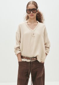 Frau mit lockigem Haar, die übergroße dunkelblaue Sonnenbrille trägt, eine lockere beige strukturierte Bluse, braune Hosen und einen genoppten Gürtel mit geflochtenem Zugband.