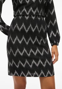 Fustă neagră cu model zigzag argintiu, material texturat și siluetă ajustată. Are mâneci lungi, terminându-se deasupra genunchiului.