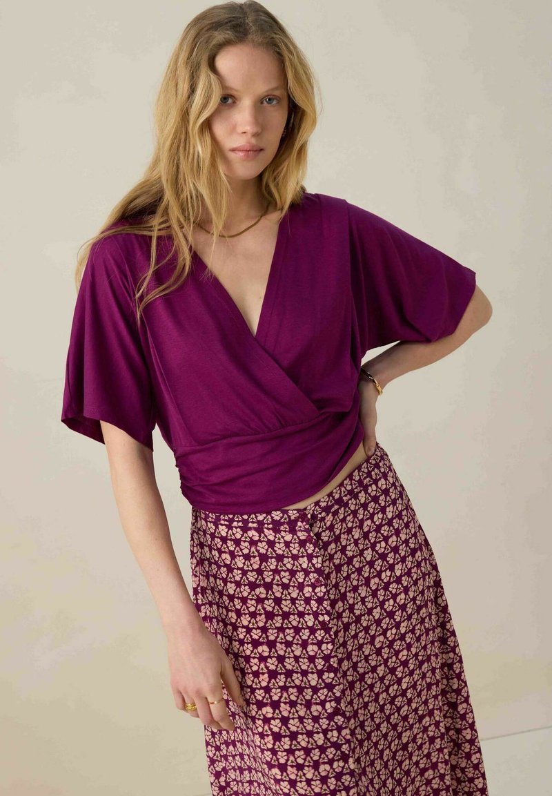 Haut à col en V et manches courtes en violet profond, associé à une jupe fluide à motifs avec des designs rose pâle sur un fond sombre.