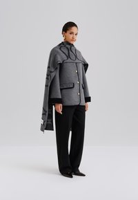 Szary płaszcz z mieszanki wełny o pudełkowym kroju, z dużym kołnierzem, złotymi guzikami i wzorzystym narzutem na ramiona. Stylizowany z czarnymi spodniami i spiczastymi butami.