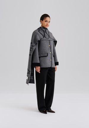 Šedý kabát z vlnenej zmesi s hranatým dizajnom, veľkým golierom, zlatými gombíkmi a vzorovaným plášťom na pleciach. Štylizovaný s čiernymi nohavicami a špicatou obuvou.