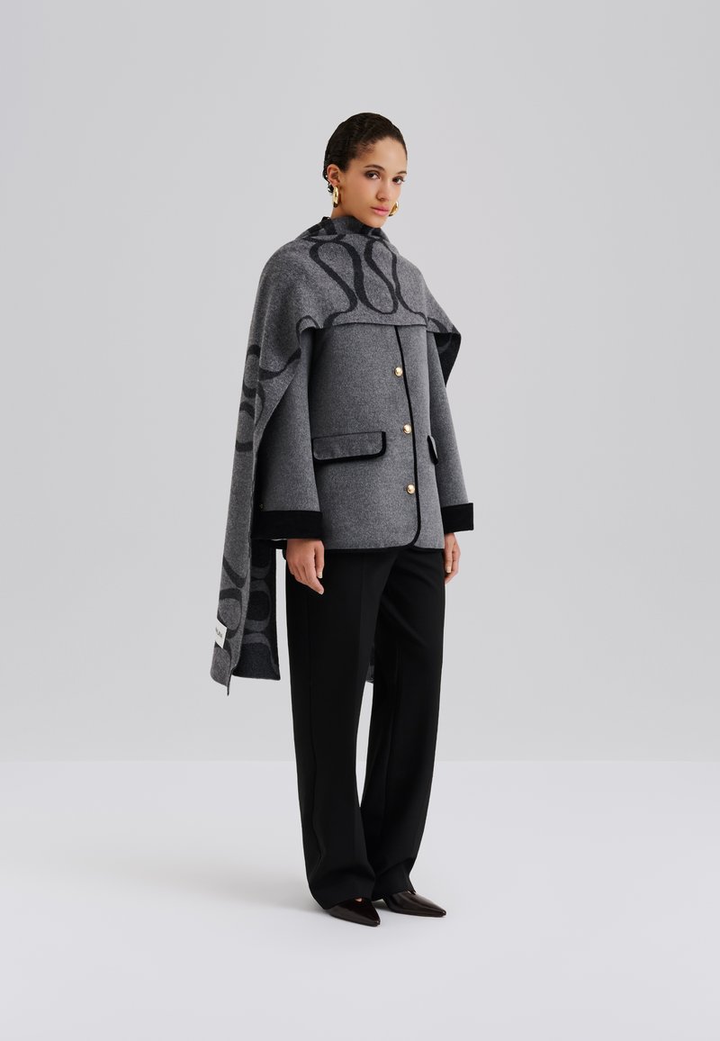 Szary płaszcz z mieszanki wełny o pudełkowym kroju, z dużym kołnierzem, złotymi guzikami i wzorzystym narzutem na ramiona. Stylizowany z czarnymi spodniami i spiczastymi butami.