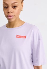 T-shirt in cotone di un leggero colore viola con un patch rosso del logo Columbia. Il design presenta una vestibilità comoda e maniche corte con scollatura a girocollo.