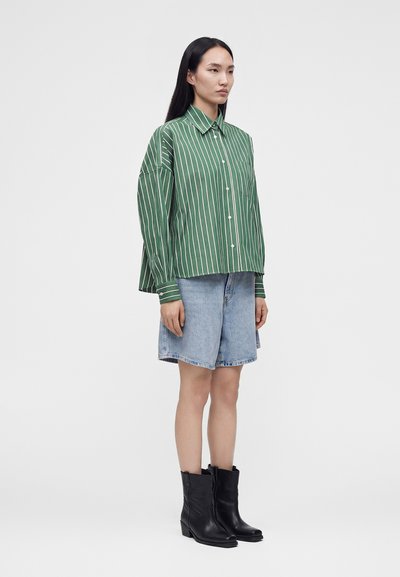 Femme debout de profil portant une chemise à rayures vertes et blanches boutonnée, un short en denim jusqu’au genou et des bottes noires mi-mollet sur fond blanc.
