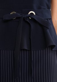 Navy sininen peplum-toppi, jossa on etutasku, hopeiset silmukat ja röyhelöhelma. Sen alla oleva tummansinisellä raidallisella hameella luodaan tekstuuri kontrasti.