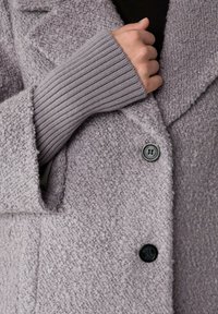 Cappotto grigio testurizzato con colletto, polsini a coste e bottoni neri. Il tessuto è morbido con un aspetto soffice e peloso.
