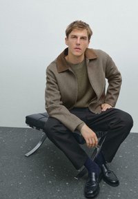 Brown wool jacket with a brown collar, green knitted sweater, black trousers, and black loafers on a black chair against a light background.