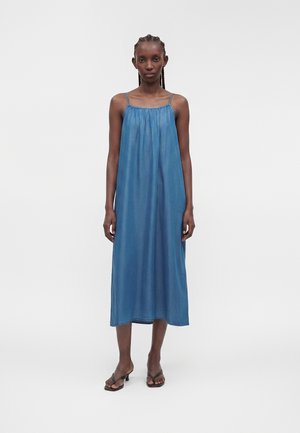 Mujer de pie vestida con un vestido midi azul sin mangas con tirantes finos y sandalias de tacón con punta cuadrada sobre un fondo blanco liso.