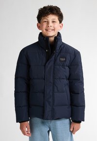 Giacca piumino blu navy con colletto alto, texture trapuntata, zip frontale e una piccola etichetta con logo sul lato sinistro superiore. Vestibilità casual.