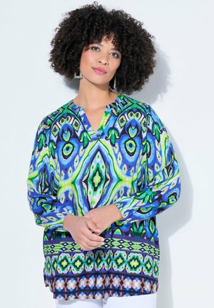 Kvinde med krøllet hår iført store firkantede øreringe og en lys mønstret bluse med lange ærmer, stående med hænderne løst foldet.