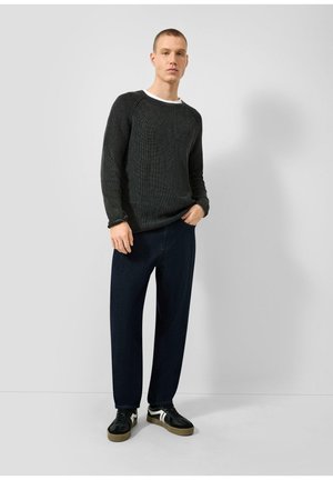 Jeune homme portant un pull à manches longues noir texturé, un pantalon foncé et des baskets noires avec des bandes blanches, debout devant un fond clair uni.