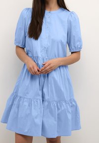 Ljusblå klänning i bomull. Har korta puffärmar, rund halsringning, knappstängning fram och ett lager-på-lager kjol. Slät textur.