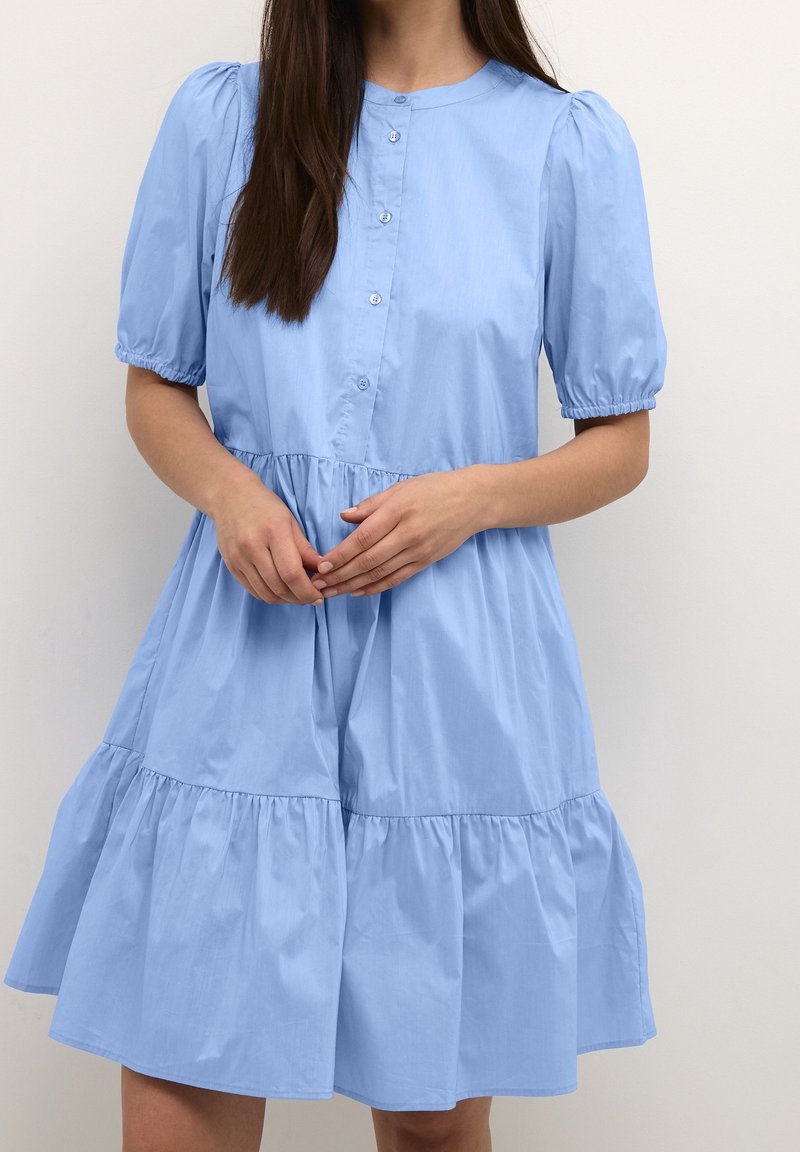 Ljusblå klänning i bomull. Har korta puffärmar, rund halsringning, knappstängning fram och ett lager-på-lager kjol. Slät textur.