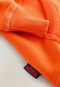 Žiarivo oranžový bavlnený sveter s rebrovanými manžetami a výraznou červenou nášivkou "Superdry" na tmavomodrom pozadí.