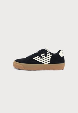 Baskets basses noires avec un logo géométrique blanc sur le côté et une semelle en gomme, montrées de profil sur un fond clair.